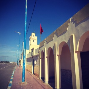 Rue et bâtiments jaunes sur la droite, un drapeau marocain flotte au vent #Off2Africa 9 Tan-Tan Tarfaya Maroc © Gilles Denizot 2016
