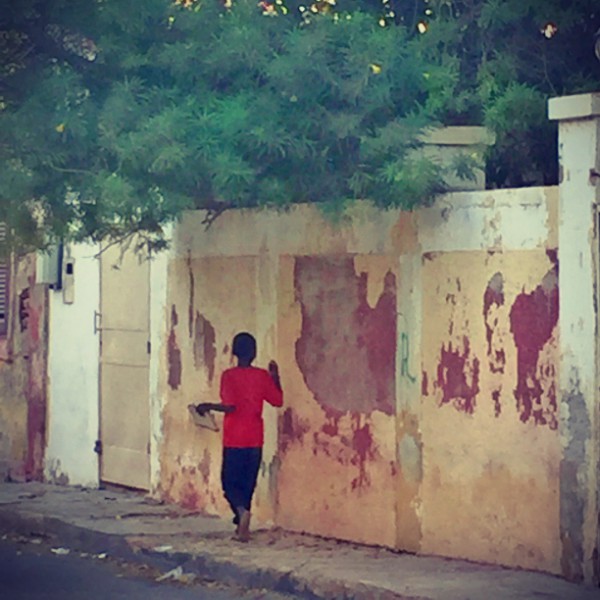 Un garçon au t-shirt rouge marche contre un mur, il tient un cahier dans la main #Off2Africa 19 Saint-Louis Sénégal © Gilles Denizot 2016