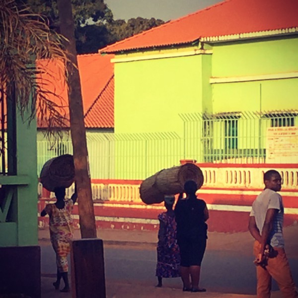 Des femmes portent de gros rouleaux sur la tête, un homme dans la rue, bâtiments verts et oranges #Off2África 50 Bissau Guinée-Bissao © Gilles Denizot 2017