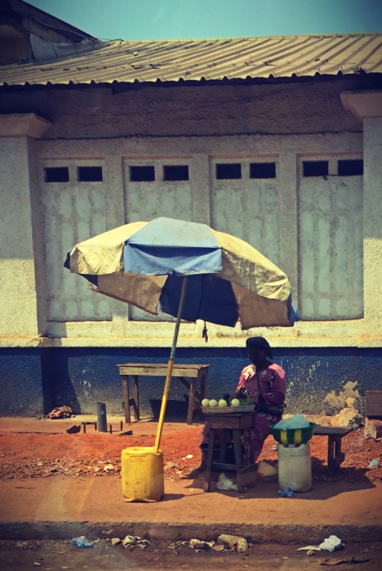 Une femme assise au bord d'une route, sous un parasol jaune et bleu #Off2Africa 69 Conakry Guinée © Gilles Denizot 2017