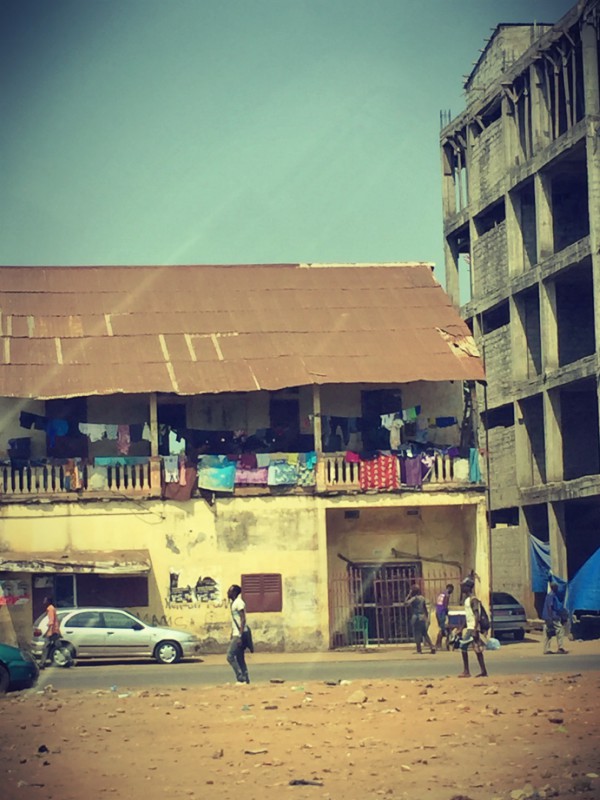 Une rue, des gens passant, une grande maison délabrée, du linge qui sèche au balcon, un bâtiment en construction sur la droite #Off2Africa 67 Conakry Guinée © Gilles Denizot 2017
