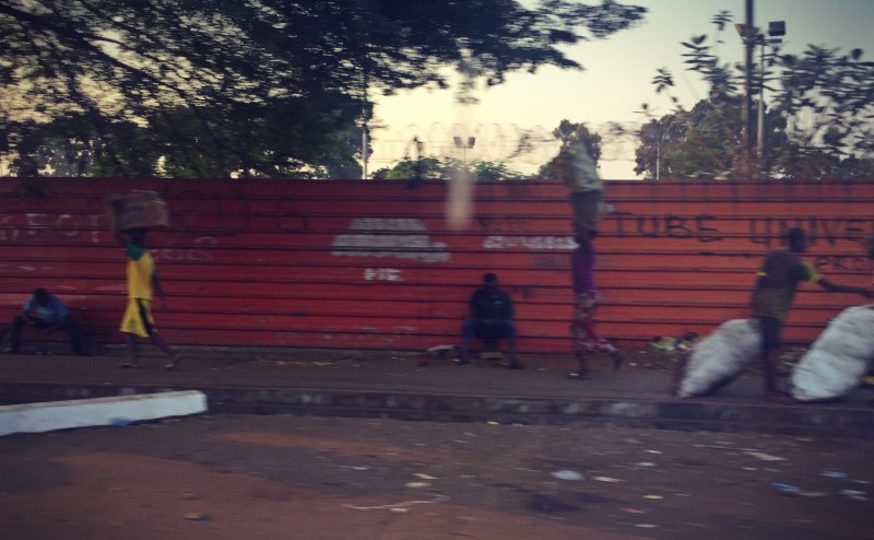 Scène de rue, des hommes et des enfants charrient des sacs devant un mur en tôle rouge #Off2Africa 65 Conakry Guinée © Gilles Denizot 2017
