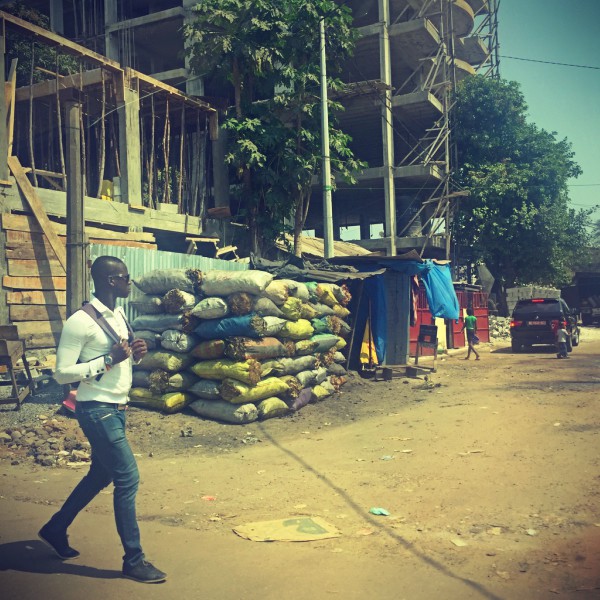 Un homme marche dans la rue, derrière lui un immeuble en construction et des sacs colorés empilés les uns sur les autres #Off2Africa 64 Conakry Guinée © Gilles Denizot 2017