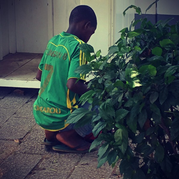Un enfant au maillot de foot vert accroupi derrière une plante #Off2Africa 68 Conakry Guinée © Gilles Denizot 2017