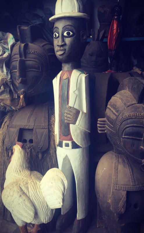 Des figurines en bois, au milieu desquelles une statuette d'un homme portant chapeau et costume blancs, et une poule blanche #Off2Africa 78 Conakry Guinée © Gilles Denizot 2017