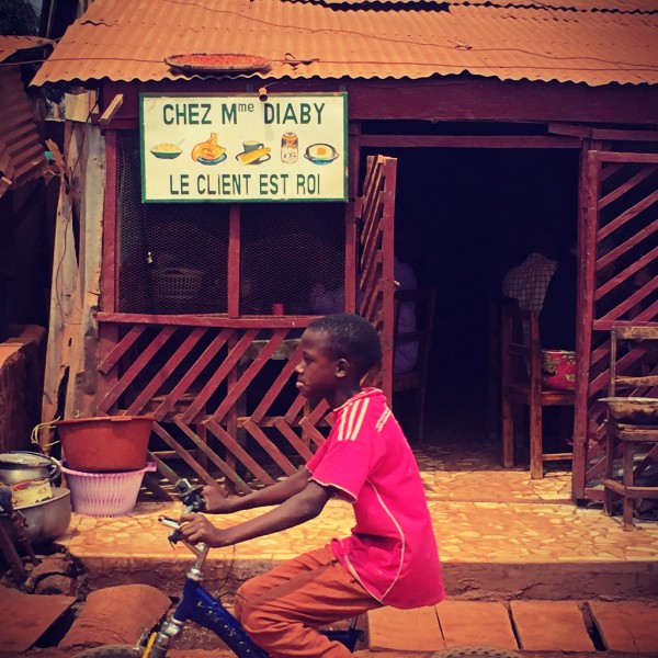Un gamin sur un vélo passe devant le restaurant de Mme Diaby #Off2Africa 83 Kankan Guinée © Gilles Denizot 2017