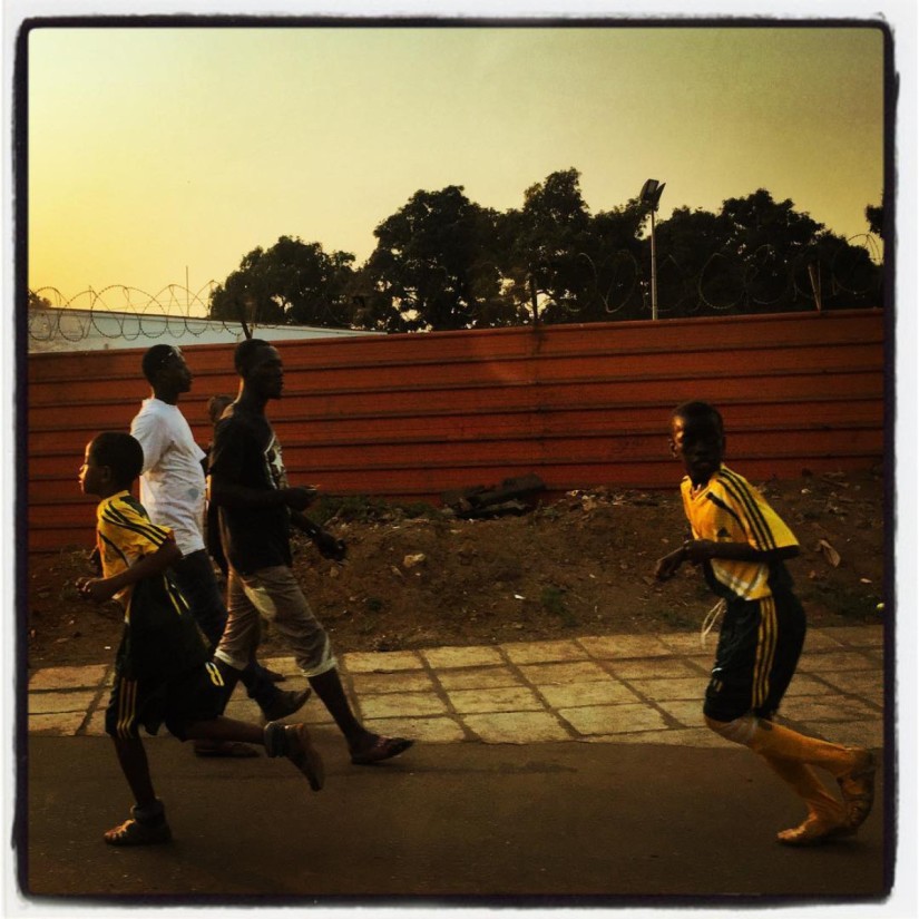 Deux garçons en tenue jaune et noir de football courent de droite à gauche, derrière eux deux hommes marchent de gauche à droite #Off2Africa 65 Conakry Guinée © Gilles Denizot 2017