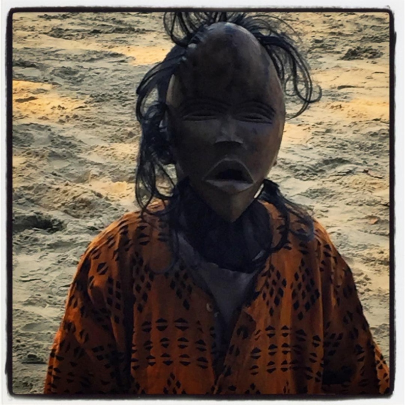 Un gamin portant un masque en bois et cheveux noirs, vêtu d'une chemise traditionnelle sur un sol de sable #Off2Africa 74 Îles de Loos Guinée © Gilles Denizot 2017
