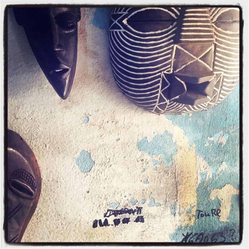 Trois masques africains en bois sont accrochés sur un mur décrépi bleu ciel et blanc, on y voit aussi quelques inscriptions au feutre noir #Off2Africa 77 Conakry Guinée © Gilles Denizot 2017