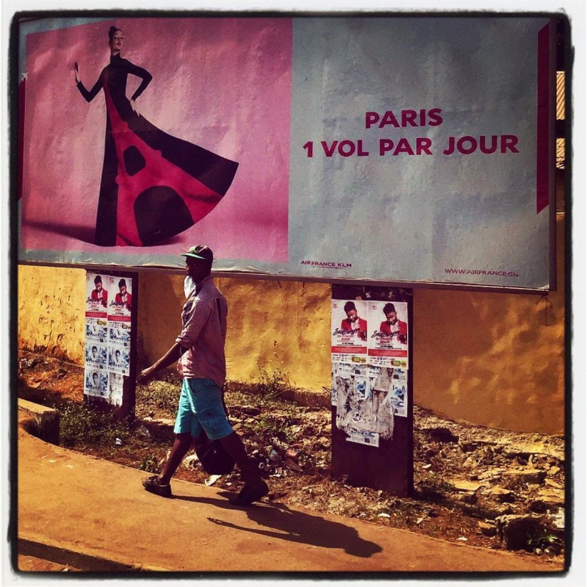 Dans une rue de Conakry, un homme vêtu d'une chemise rose marche sous un panneau publicitaire rose d'Air France #Off2Africa 80 Conakry Guinée © Gilles Denizot 2017