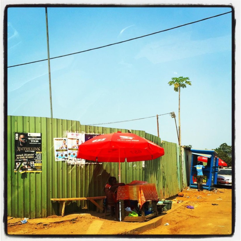 Dans une rue ensablée, un mur en tôle métallique verte, un arbre et un stand au parasol rouge #Off2Africa 69 Conakry Guinée © Gilles Denizot 2017