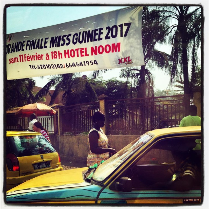 Dans une rue de Conakry, une banderole annonce la grande finale de Miss Guinée 2017, deux taxis et deux femmes #Off2Africa 78 Conakry Guinée © Gilles Denizot 2017