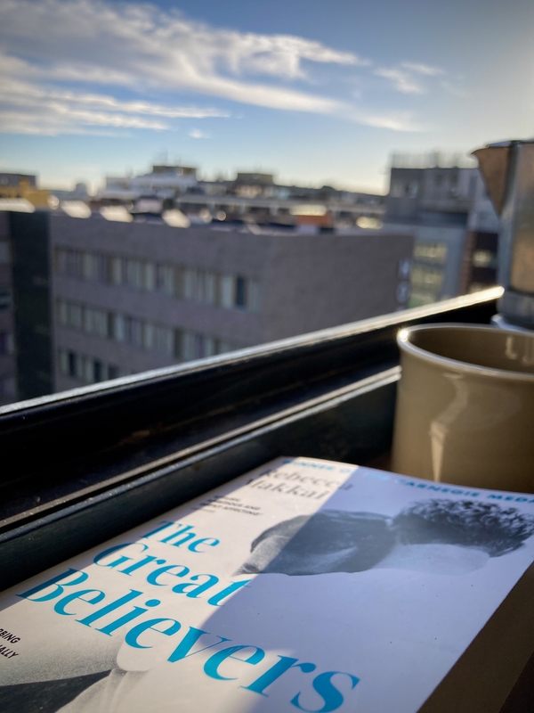 Le livre The Great Believers, une tasse de café et une cafetière. Au loin, la vue depuis la fenêtre ouverte © Gilles Denizot 2023