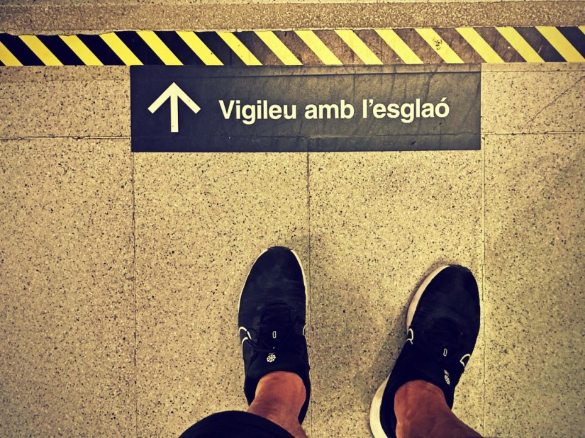 Vue d'en haut sur chaussures de sport noires et un avertissement en catalan. Lignes obliques jaunes et noires. #HolaBarcelona juillet 2023 © Gilles Denizot