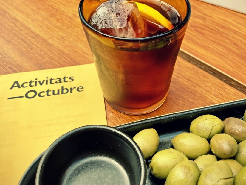 Sur une table en bois, un verre de vermouth, des olives vertes et un feuillet Activitats Octubre jaune #HolaBarcelona octobre 2022 © Gilles Denizot